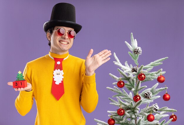 Young man in yellow turtleneck and glasses wearing black hat and funny tie holding cubes with new year date 