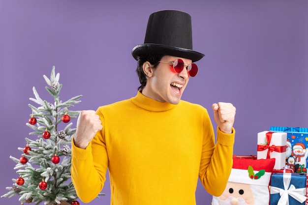 黄色いタートルネックと黒い帽子をかぶった眼鏡の若い男は、クリスマスツリーの横に立って紫色の壁の上に立って幸せで興奮した握りこぶし