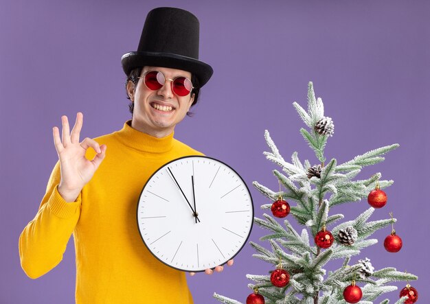 黄色のタートルネックと眼鏡をかけた若い男が壁掛け時計を持ってカメラを見て笑って、紫色の背景の上のクリスマスツリーの横に立っているOKサインを示しています