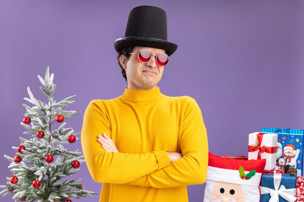 Молодой человек в желтой водолазке и очках в черной шляпе смотрит в камеру, недовольный, скрестив руки на груди, стоит рядом с елкой и представляет подарки на фиолетовом фоне