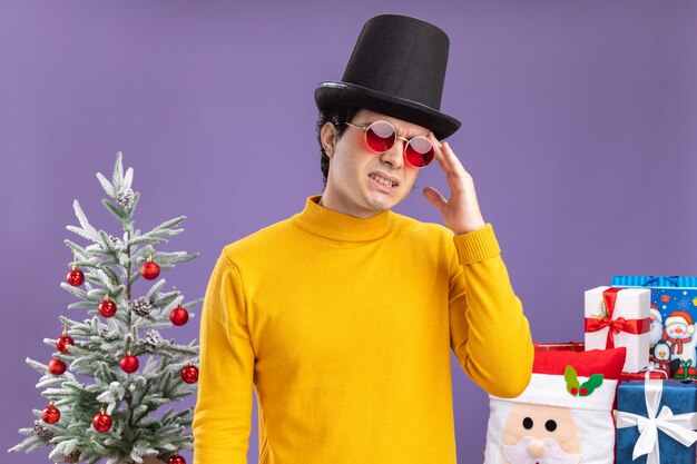 Молодой человек в желтой водолазке и очках в черной шляпе выглядит нездоровым, трогает свою голову с головной болью, стоит рядом с елкой и преподносит подарки через фиолетовую стену