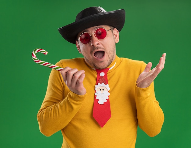 노란색 터틀넥과 녹색 벽 위에 사탕 지팡이를 들고 검은 모자에 재미있는 빨간 넥타이와 안경에 젊은 남자가 행복하고 흥분 서