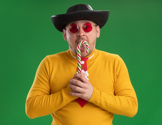Young man in yellow turtleneck and glasses with funny red tie in black hat holding candy cane  surprised standing over green wall