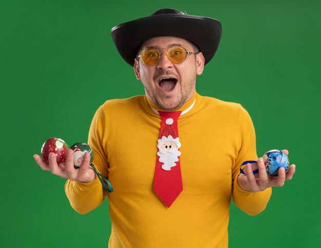노란색 터틀넥과 녹색 벽 위에 크리스마스 트리 행복하고 흥분 서 장난감을 들고 재미있는 빨간 넥타이와 안경에 젊은 남자