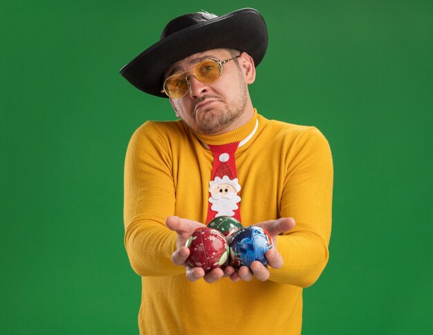 노란색 터틀넥과 녹색 벽 위에 서있는 슬픈 표정으로 크리스마스 트리 장난감을 들고 재미있는 빨간 넥타이와 안경에 젊은 남자