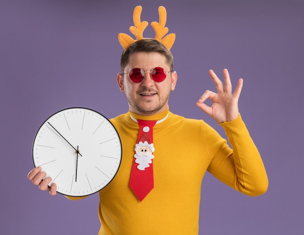 黄色のタートルネックと赤い眼鏡の若い男は、紫色の背景の上に立っているOKのサインを示しているカメラの笑顔を見て壁時計を保持している頭に鹿の角を持つ面白い赤いネクタイと縁を身に着けています