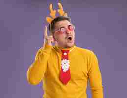 Free photo young man in yellow turtleneck and red glasses wearing funny red tie and rim with deer horns on head looking aside surprised showing index finger having new great idea standing over purple background