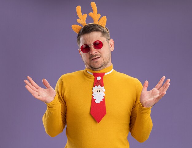 young man in yellow turtleneck and red glasses wearing funny red tie and rim with deer horns on head looking at camera confused shrugging shoulders standing over purple background