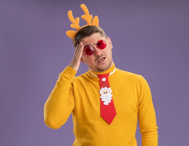 Young man in yellow turtleneck and red glasses wearing funny red tie and rim with deer horns on head looking confused and very anxious standing over purple background