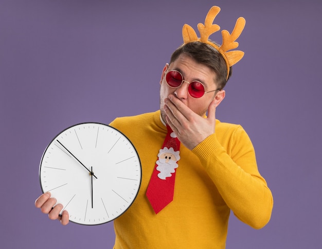 Free photo young man in yellow turtleneck and red glasses wearing funny red tie and rim with deer horns on head looking at it amazed and surprised standing over purple background