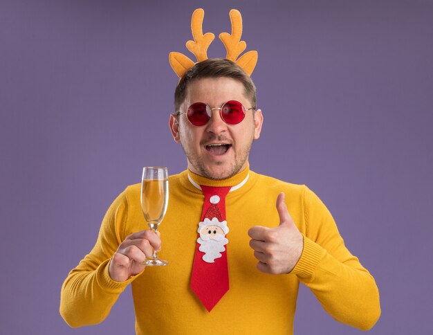黄色いタートルネックと赤い眼鏡をかけた若い男が面白い赤いネクタイと縁を身に着けて、鹿の角がシャンパンのグラスを持って幸せで陽気な笑顔を見せて親指を立てて立っている