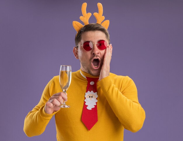 Молодой человек в желтой водолазке и красных очках, в забавном красном галстуке и оправе с оленьими рогами, держит бокал шампанского, глядя в камеру, изумленный и удивленный, стоя на фиолетовом фоне