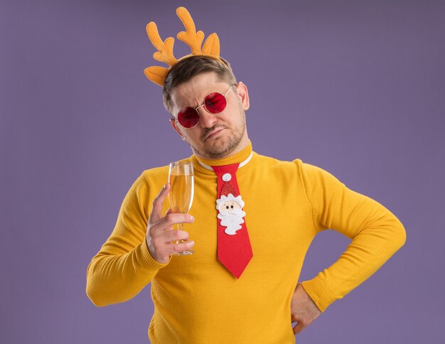 Молодой человек в желтой водолазке и красных очках в забавном красном галстуке и оправе с оленьими рогами держит бокал шампанского и недовольно смотрит на него, стоя на фиолетовом фоне