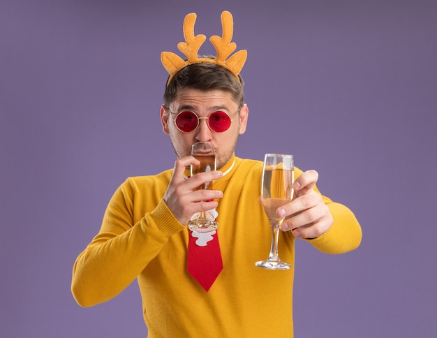 Free photo young man in yellow turtleneck and red glasses wearing funny red tie and rim with deer horns holding two glasses of champagne happy and positive standing over purple background