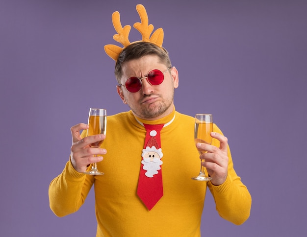 Free photo young man in yellow turtleneck and red glasses wearing funny red tie and rim with deer horns holding two glasses of champagne looking confused and displeased standing over purple background