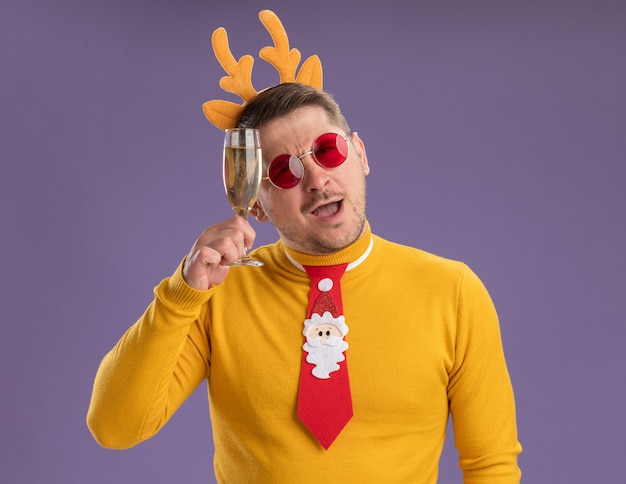 Free photo young man in yellow turtleneck and red glasses wearing funny red tie and rim with deer horns looking at camera happy and positive holding glass of champagne standing over purple background