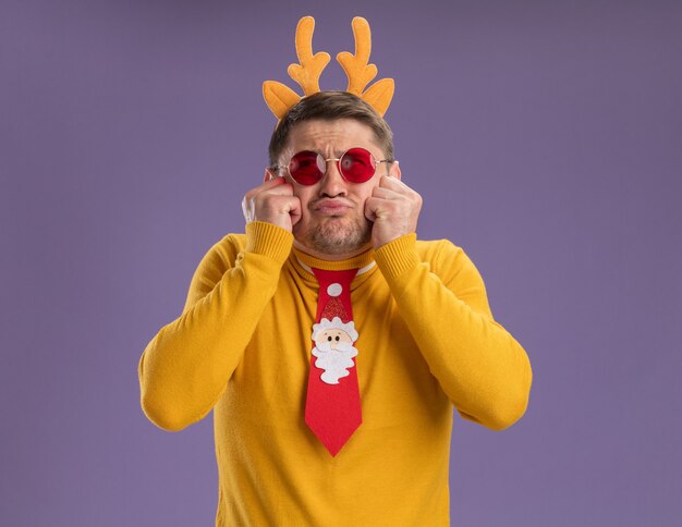 young man in yellow turtleneck and red glasses wearing funny red tie and rim with deer horns looking up confused and disappointed standing over purple background