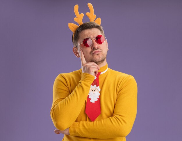 Young man in yellow turtleneck and red glasses wearing funny red tie and rim with deer horns looking up with pensive expression thinking standing over purple background