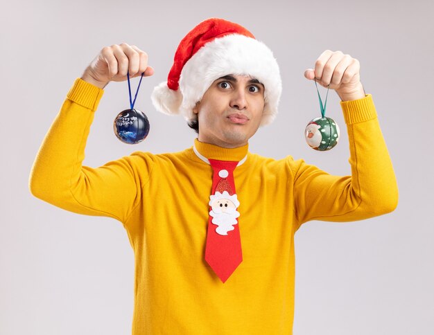 Young man in yellow turtleneck and santa hat holding christmas balls  confused standing over white wall