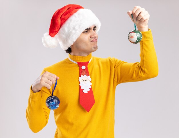 Young man in yellow turtleneck and santa hat holding christmas balls looking at them confused having doubts standing over white wall