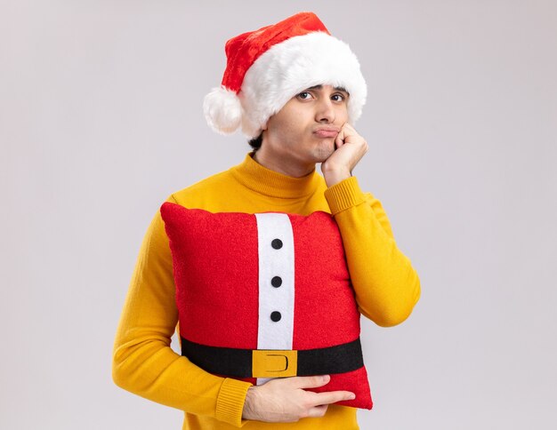 Молодой человек в желтой водолазке и шляпе санта-клауса, держащий рождественскую подушку, уставший и скучающий, глядя в камеру, стоит на белом фоне