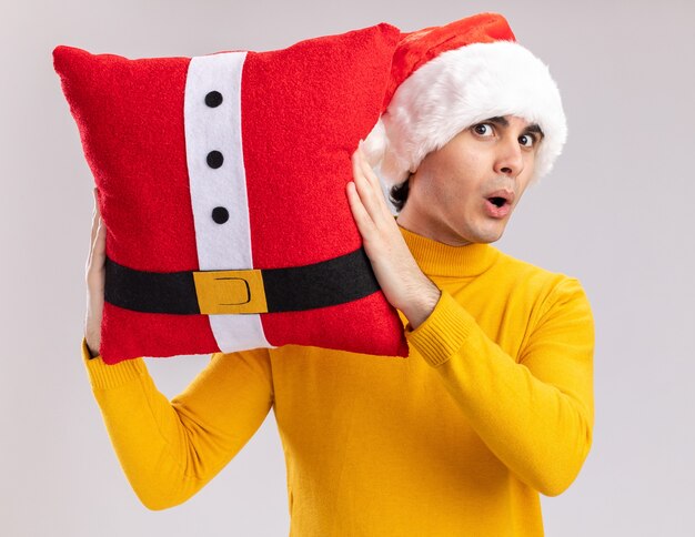 Young man in yellow turtleneck and santa hat holding christmas pillow  surprised and amazed standing over white wall