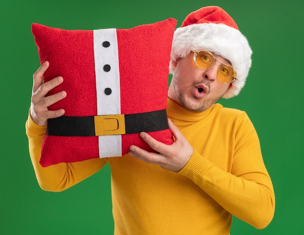 Young man in yellow turtleneck and santa hat wearing glasses holding red funny pillow looking at camera happy and surprised standing over green background