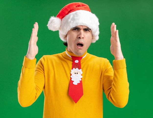 Young man in yellow turtleneck and santa hat with funny tie  amazed showing large size gesture with hands , measure symbol standing over green wall