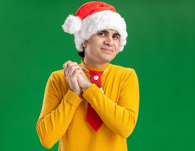Young man in yellow turtleneck and santa hat with funny tie holding hands together happy and cheerful waiting for surprise standing over green background