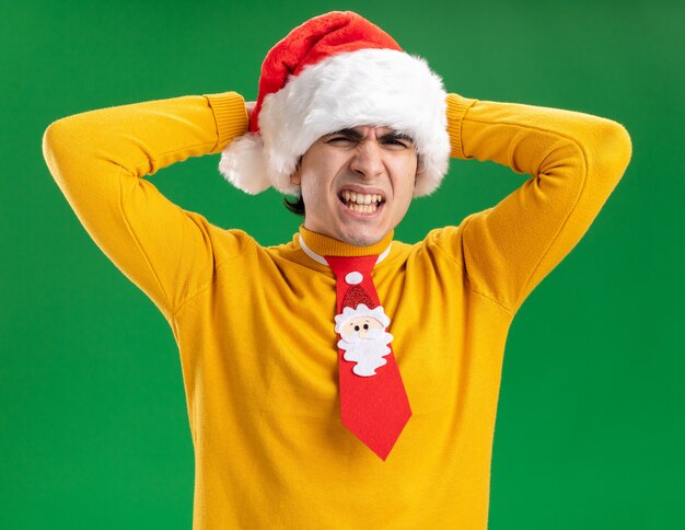 Young man in yellow turtleneck and santa hat with funny tie looking annoyed and irritated with hands on his head standing over green background