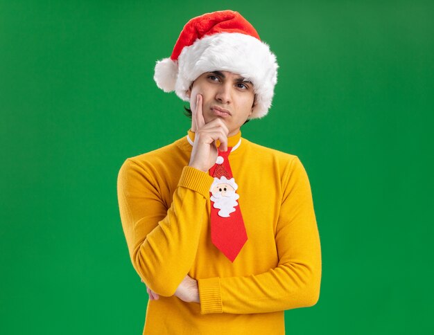 Young man in yellow turtleneck and santa hat with funny tie looking aside with hand on chin with pensive expression thinking standing over green background