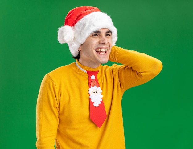 Young man in yellow turtleneck and santa hat with funny tie looking aside with happy face smiling cheerfully standing over green background