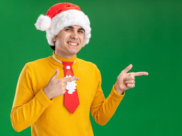 Young man in yellow turtleneck and santa hat with funny tie looking at camera smiling cheerfully pointing with index fingers to the side standing over green background