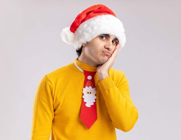 Young man in yellow turtleneck and santa hat with funny tie looking at camera tired and bored standing over white background