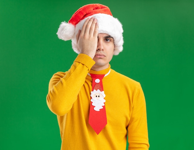 Young man in yellow turtleneck and santa hat with funny tie looking at camera wirth serious face covering one eye with hand standing over green background