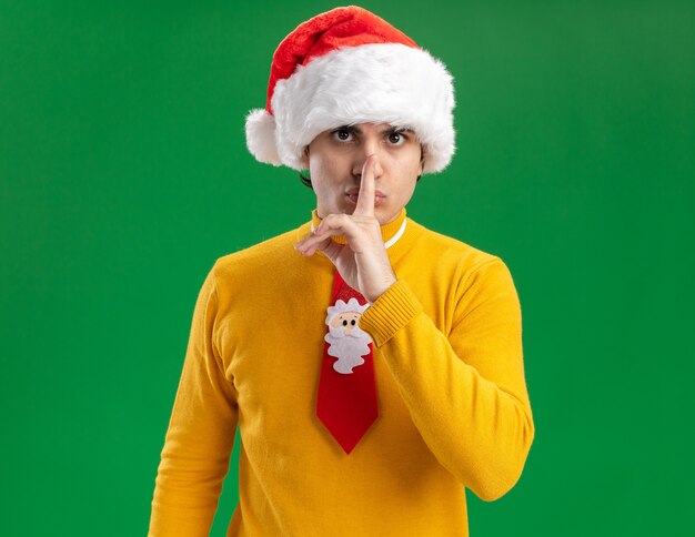 Young man in yellow turtleneck and santa hat with funny tie looking at camera with serious face making silence gesture with finger on lips standing over green background