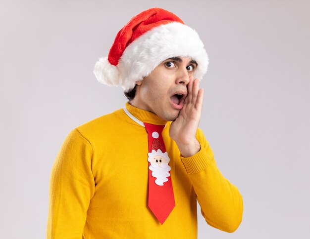 Young man in yellow turtleneck and santa hat with funny tie telling a secret with arm near mouth looking surprised standing over white background