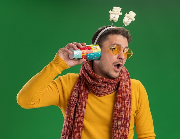 Foto gratuita giovane uomo in dolcevita giallo con sciarpa calda e occhiali da indossare bordo divertente sulla testa che tiene tazza colorata sopra l'orecchio cercando di ascoltare i pettegolezzi in piedi sopra la parete verde