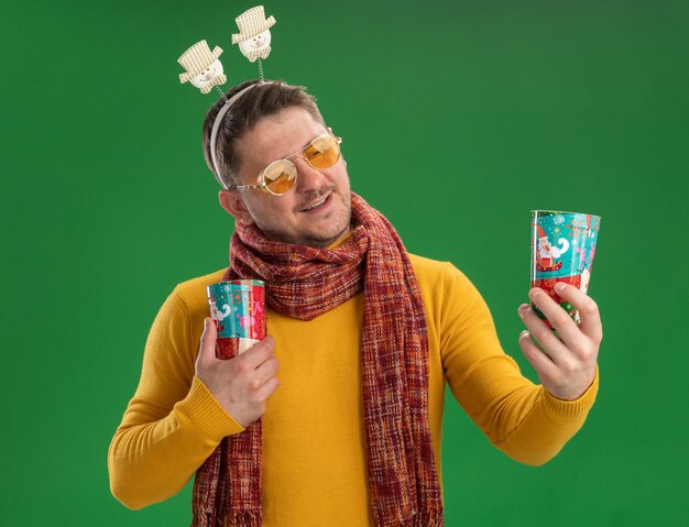 Young man in yellow turtleneck with warm scarf and glasses wearing funny rim on head holding colorful cups looking at them with smile on face standing over green wall