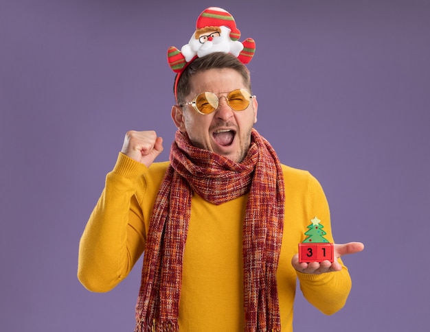 Молодой человек в желтой водолазке с теплым шарфом и очками в забавной оправе с дедом морозом на голове показывает игрушечные кубики с новогодней датой, сжимая кулаки, сумасшедший счастливый, стоящий над фиолетовой стеной