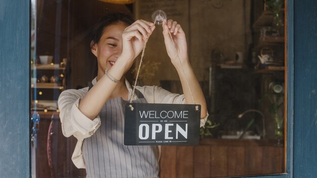 Young manager girl changing a sign from closed to open sign on door cafe looking outside waiting for clients after lockdown.