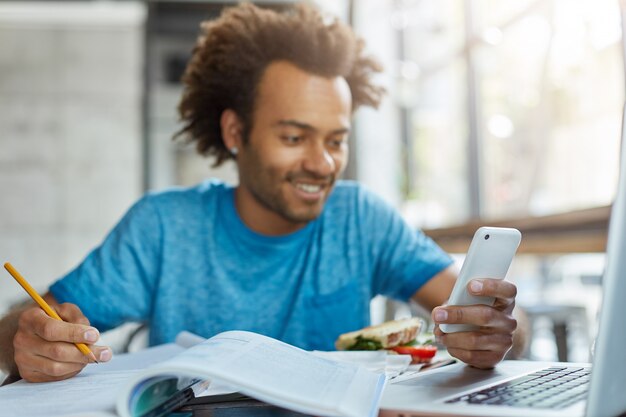 Young manager male sitting indoors writing notes using modern telephone and laptop for solving working issues.