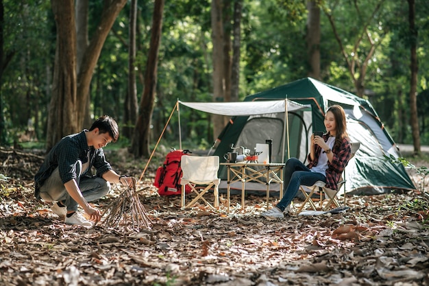 Young mand picking branches and put them together, He preparing preparing a pile of firewood for fire camping on night and pretty girl friend sitting at front of camping tent