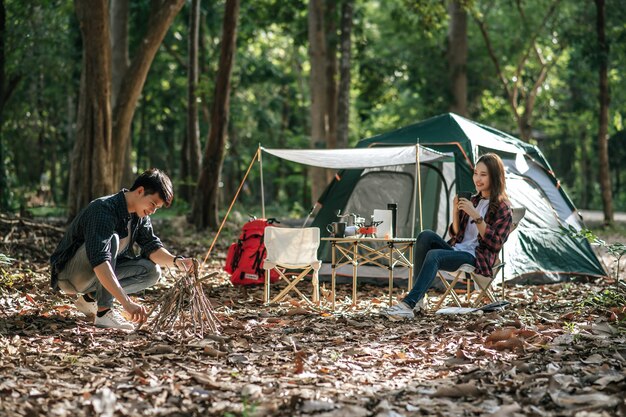 若いマンドは枝を選んでそれらをまとめました、彼は夜の火のキャンプのために薪の山を準備する準備をしていて、かわいいガールフレンドはキャンプテントの前に座っています