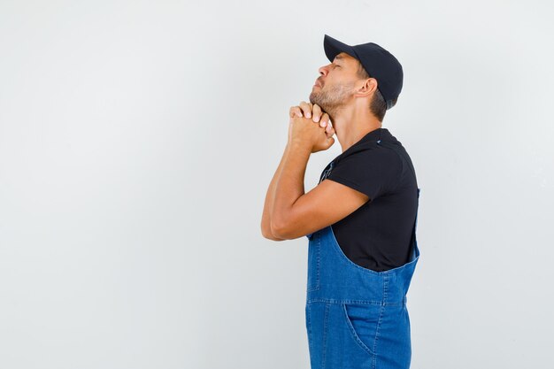 Young mechanic clasping hands in praying gesture in uniform and looking hopeful. .