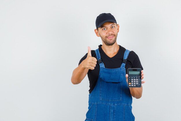 Young mechanic in uniform holding calculator with thumb up and looking cheerful , front view.