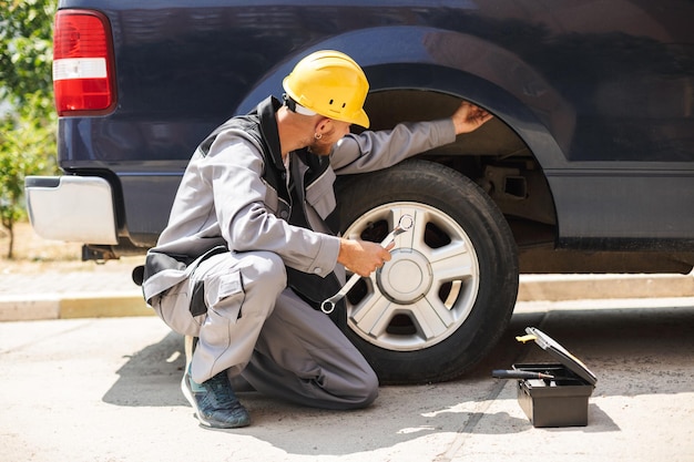 Young mechanic in work clothes and yellow hardhat using wrench for changing car wheel outdoor