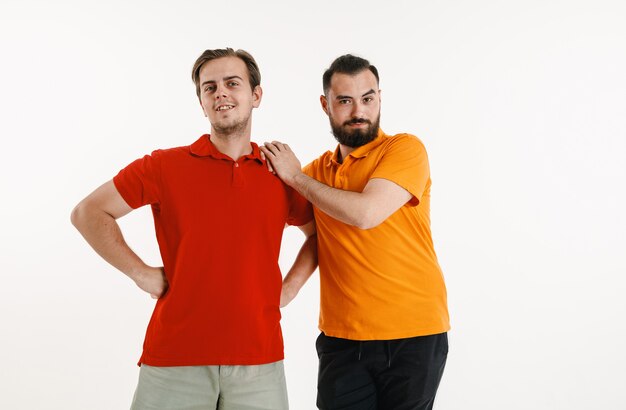 Young men weared in LGBT flag colors isolated on white wall. Caucasian male models in bright shirts. Look happy, smiling and hugging. LGBT pride, human rights and choice concept.