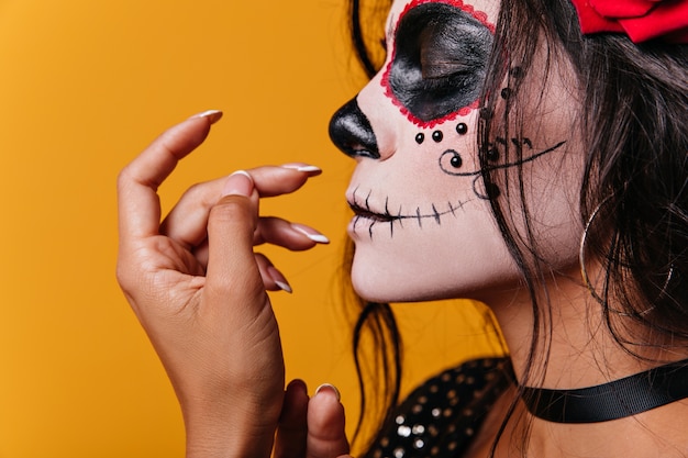 Молодая мексиканская девушка с розами в волосах и изображением черепа на лице мило позирует с закрытыми глазами
