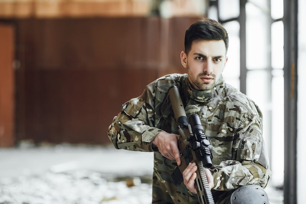 A young military soldier sits with a big rifle in his hands, near the window of a collapsed building